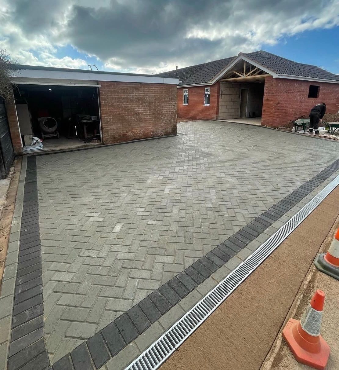 Large grey block paving driveway with linear drainage channels beside brick outbuildings